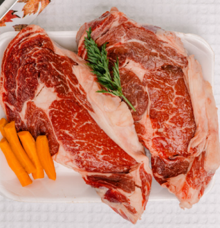 Two thick raw Ribeye Steaks on a white tray, garnished with carrots and rosemary or similar herb.