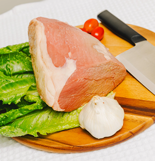 A large cut of Whole Beef on a wooden cutting board with lettuce, cherry tomatoes, a bulb of garlic, and a knife