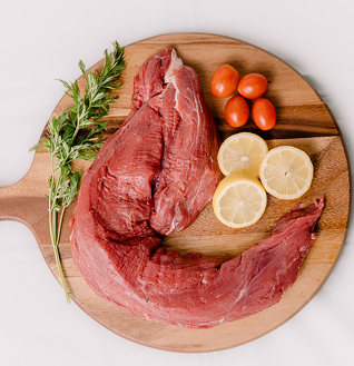 Whole raw Beef Tenderloin on a wooden cutting board, garnished with rosemary, cherry tomatoes, and lemon slices.